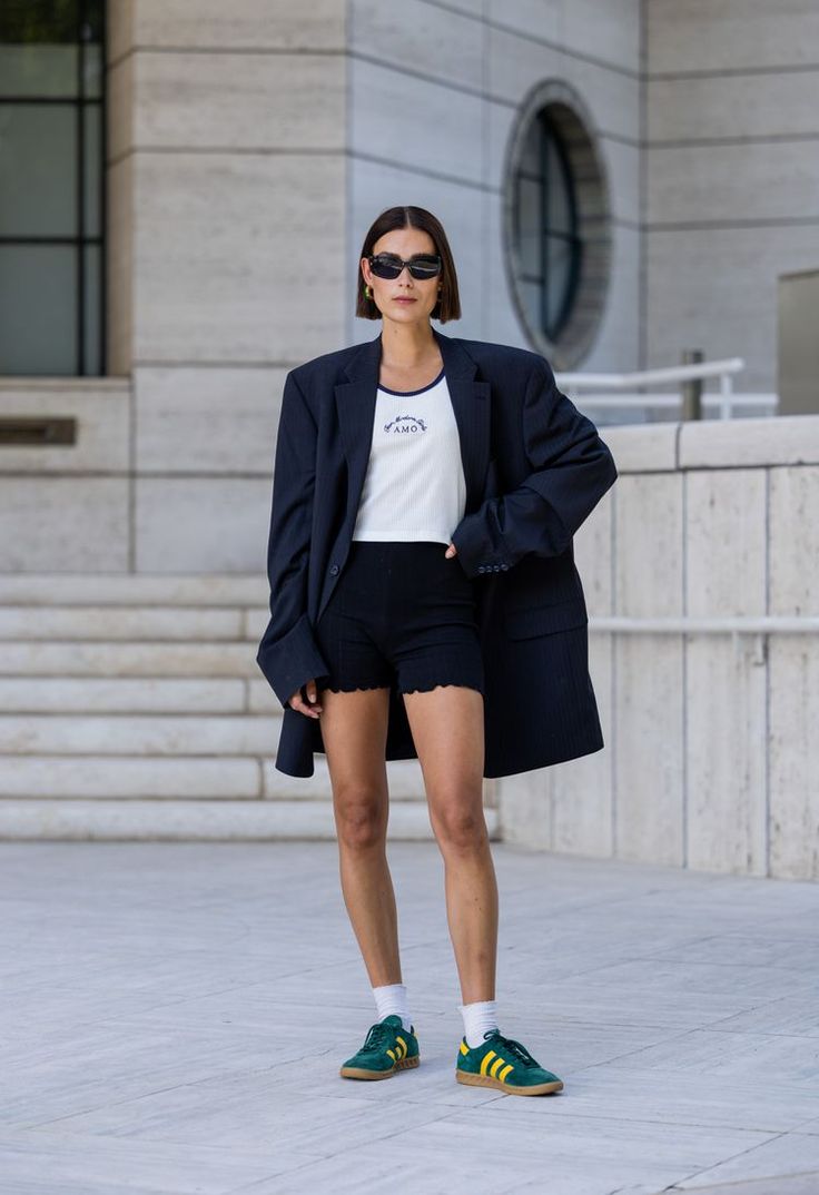 Mulher usando blazer oversized preto com shorts e tênis verde, em ambiente urbano.