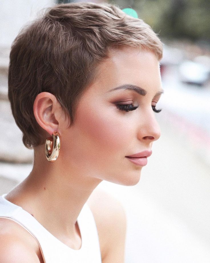 Mulher com corte pixie cut, perfil delicado e maquiagem natural, usando brincos dourados.