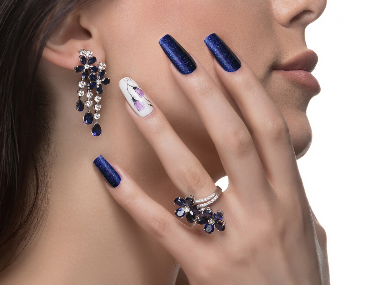 Mão feminina com unhas azul marinho cintilantes e unha decorada com flores roxas, ao lado de rosto e brincos combinando.