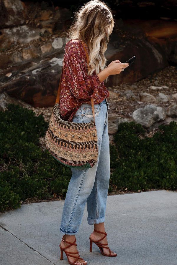 Mulher com blusa estampada estilo boho, jeans claros e sandálias de salto, carregando bolsa de palha em ambiente externo