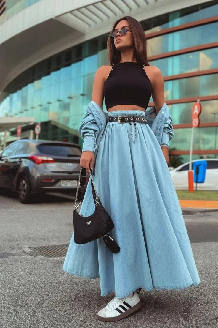Mulher usando saia godê jeans azul clara com top preto e tênis branco, em ambiente urbano moderno
