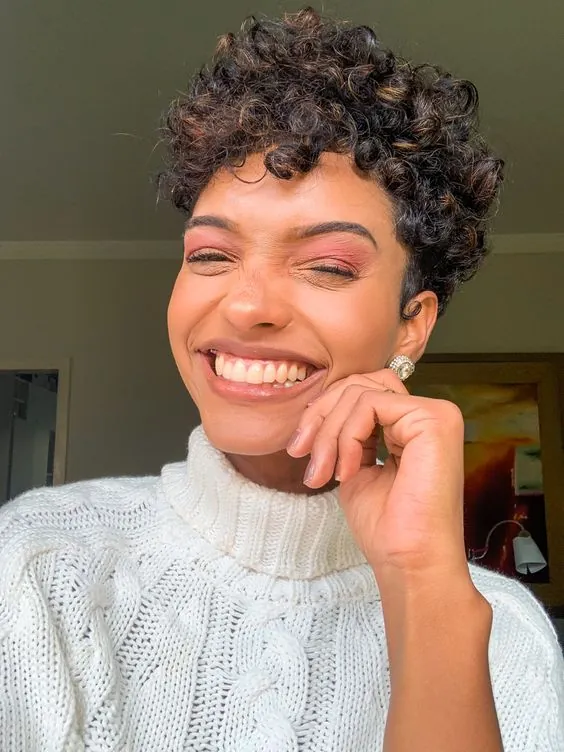 Mulher sorridente com corte pixie cacheado e suéter branco de gola alta.