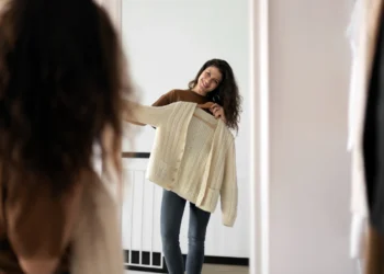 Mulher sorridente segurando uma blusa de tricô creme em frente ao espelho, avaliando a roupa para decidir se combina com seu estilo.