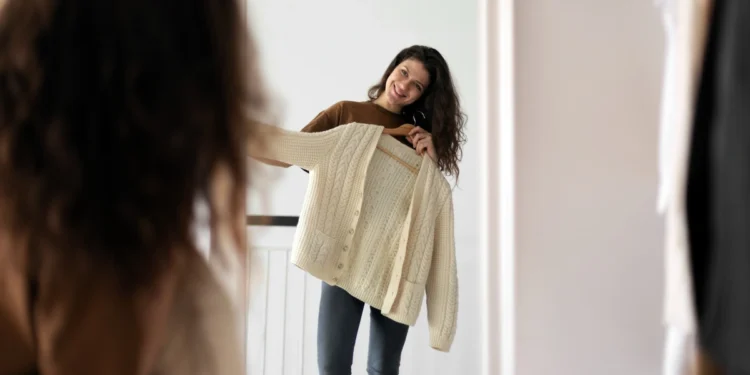 Mulher sorridente segurando uma blusa de tricô creme em frente ao espelho, avaliando a roupa para decidir se combina com seu estilo.