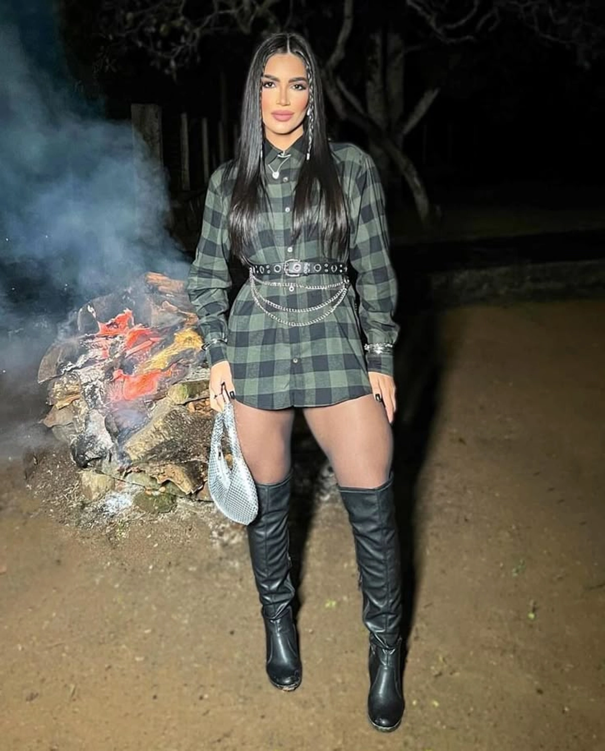 Mulher com vestido camisa xadrez preta e verde, cinto preto com correntes, botas de cano alto pretas e bolsa prata, estilo Country para São João 2025.