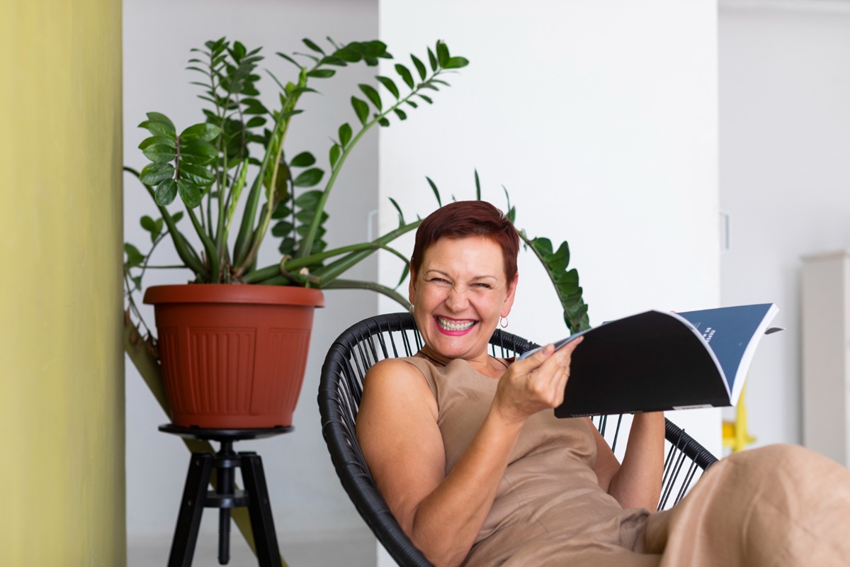 Mulher sorrindo, relaxando em cadeira, lendo livro, com planta ao fundo.