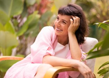 Mulher sorrindo, com mão no rosto, vestindo roupão rosa em ambiente externo.