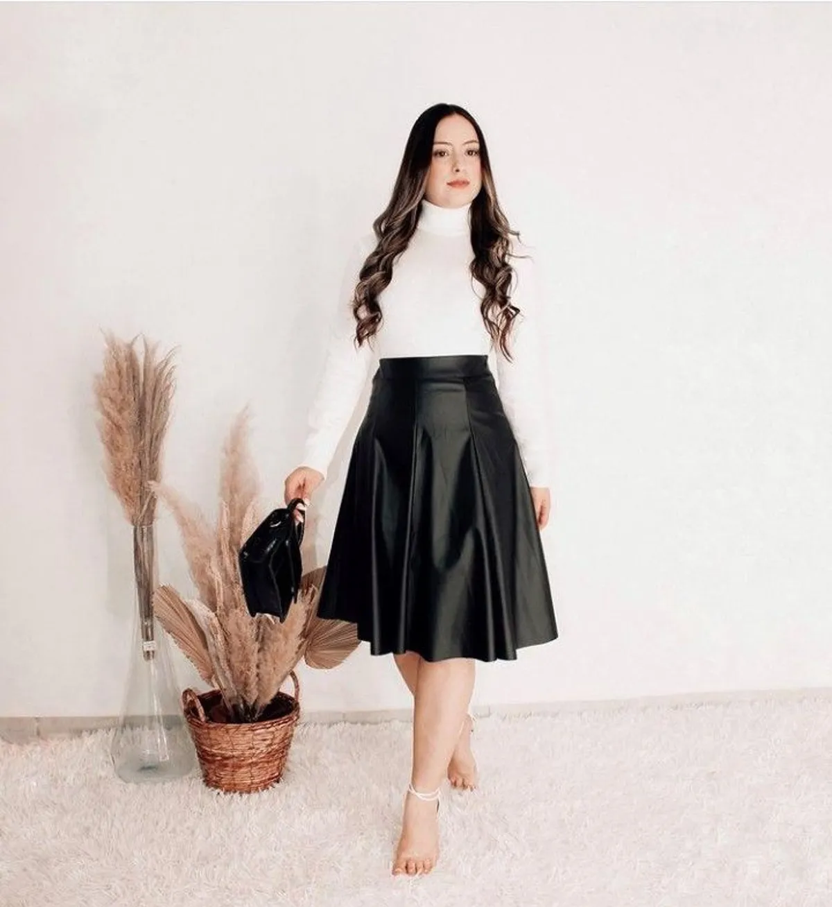 Mulher usando blusa de manga longa branca e saia godê de couro preto, com cabelo solto e segurando uma bolsa preta, posando sobre um fundo neutro.