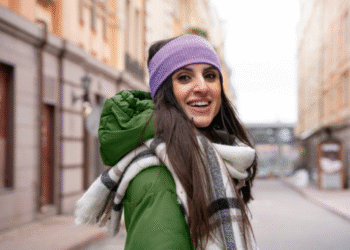 Mulher sorridente vestindo jaqueta verde, cachecol xadrez e faixa roxa na cabeça em rua urbana.