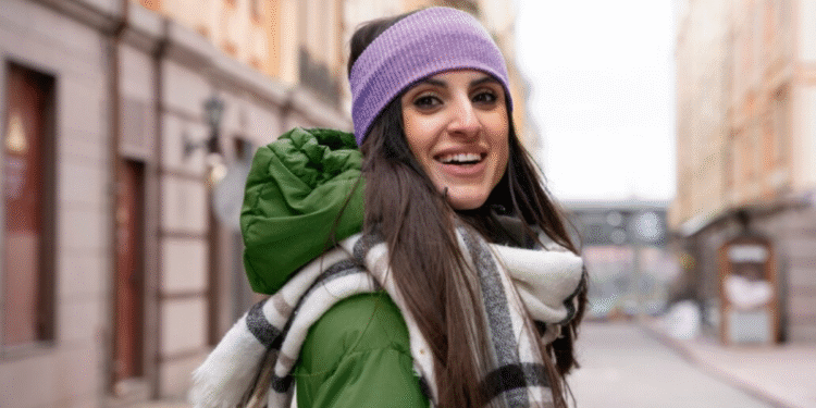 Mulher sorridente vestindo jaqueta verde, cachecol xadrez e faixa roxa na cabeça em rua urbana.