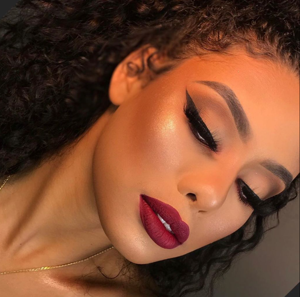 Mulher com pele escura, cabelo liso e batom vermelho vibrante, com maquiagem glamourosa.