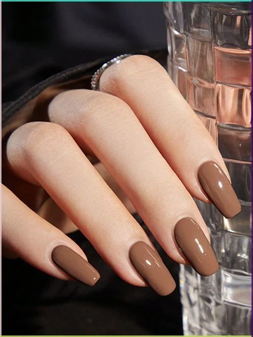 Close-up das unhas pintadas com o tom Mocha Mousse, estilo coffin, com um anel prateado e fundo escuro.
