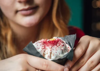 Jovem segurando um doce embrulhado em papel preto com cobertura branca e confeitos vermelhos