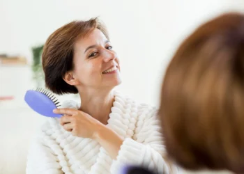 Mulher escovando o cabelo em um espelho durante a rotina de cuidados capilares na menopausa.