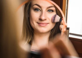 Mulher sorridente aplicando maquiagem leve e natural com blush no rosto.