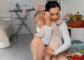 Mãe exausta sentada no quarto segurando seu bebê no colo, representando os desafios da maternidade e trabalho.