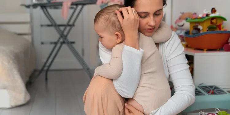 Maternidade e trabalho: as dificuldades do dia a dia precisam ser discutidas 1 Mãe exausta sentada no quarto segurando seu bebê no colo, representando os desafios da maternidade e trabalho.