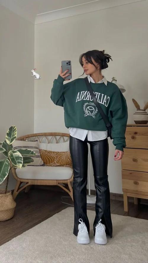 Mulher usando suéter oversized verde e calça de couro preta, com tênis branco e ambiente descontraído com móveis modernos e plantas.