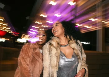 Duas mulheres sorrindo e se divertindo sob luzes coloridas à noite, usando roupas elegantes e casacos de pelúcia.