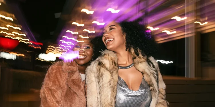Duas mulheres sorrindo e se divertindo sob luzes coloridas à noite, usando roupas elegantes e casacos de pelúcia.