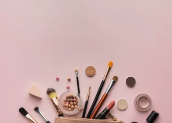 Produtos de maquiagem para iniciantes dispostos sobre uma superfície rosa, incluindo pincéis, blush, batom e outros itens essenciais.