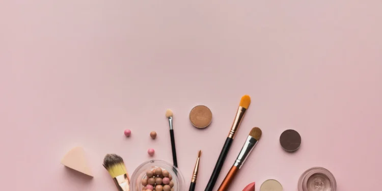 Produtos de maquiagem para iniciantes dispostos sobre uma superfície rosa, incluindo pincéis, blush, batom e outros itens essenciais.
