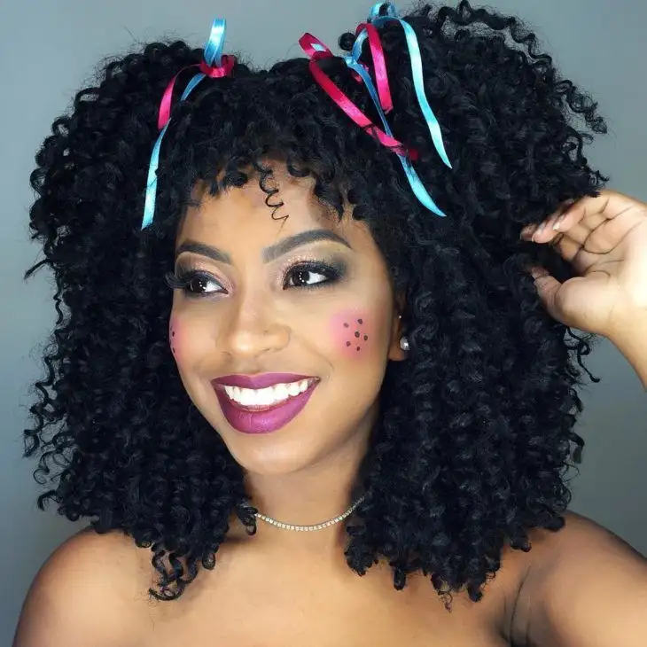 Mulher sorridente com cabelo crespo volumoso em maria-chiquinha decorado com fitas coloridas.