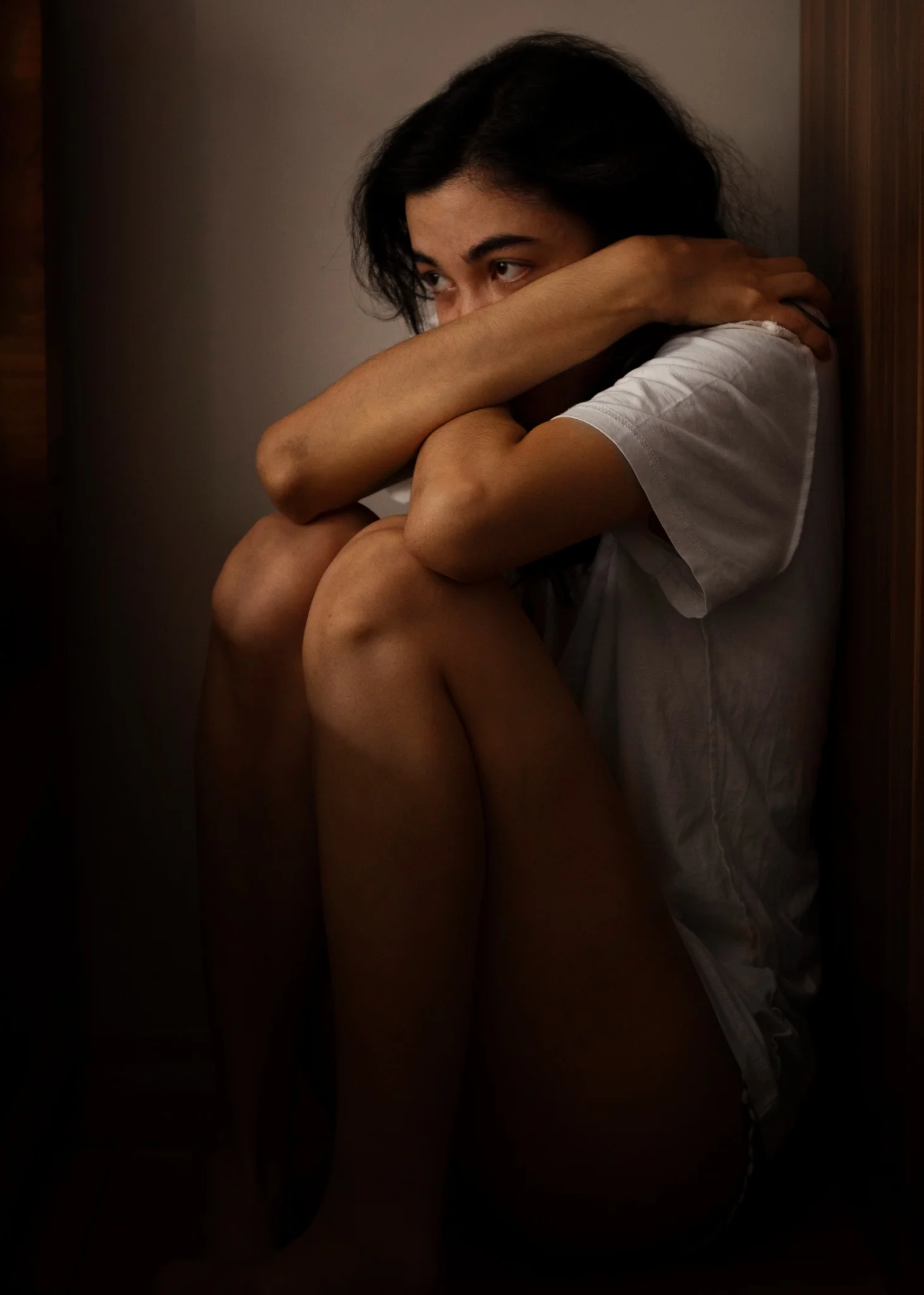 Mulher jovem, com expressão de angústia, sentada no canto de um ambiente, com os braços abraçados aos joelhos, refletindo sinais de sofrimento emocional devido à violência psicológica.