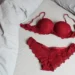 Conjunto de lingerie vermelha sobre uma cama com lençóis brancos, mostrando o sutiã e a calcinha de renda delicada.