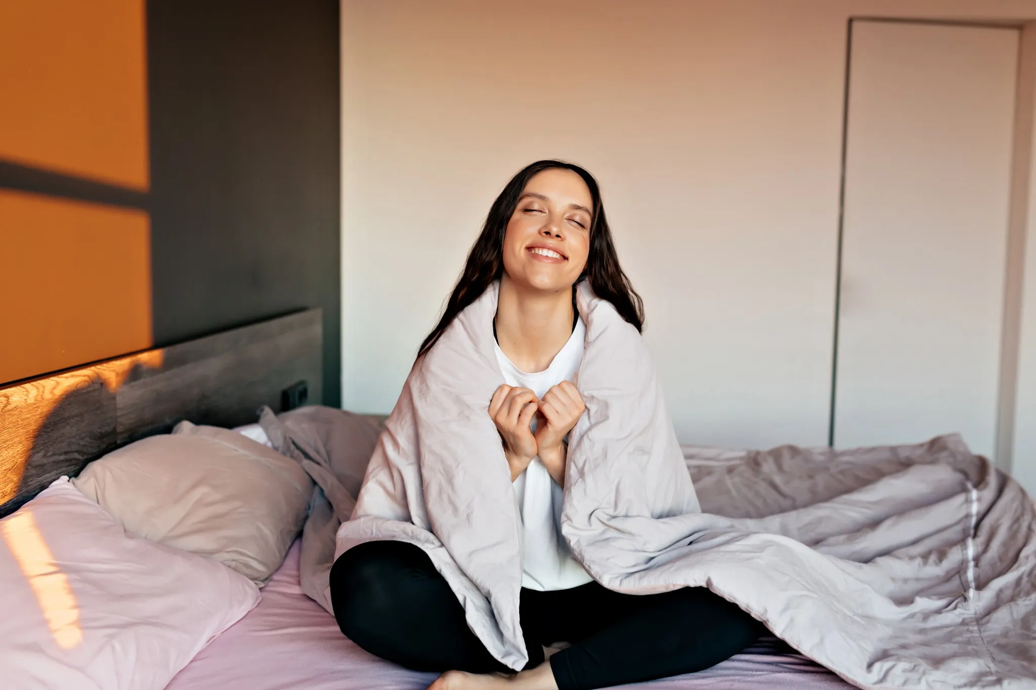 Mulher sorrindo e relaxando na cama ao amanhecer, envolta em um cobertor. 