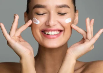 Mulher sorrindo aplicando creme facial em sua pele, representando uma rotina de cuidados com a pele.