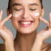 Cuidados com a pele: aprenda a rotina ideal para o seu tipo de pele! 13 Mulher sorrindo aplicando creme facial em sua pele, representando uma rotina de cuidados com a pele.