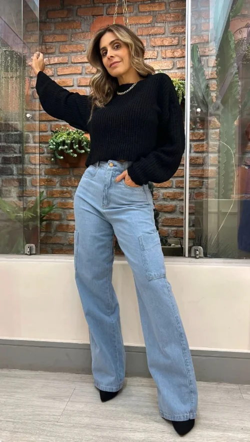 Mulher com blusa de tricô preta e calça wide leg azul, pose confiante em ambiente interno com paredes de tijolos expostos e plantas ao fundo.