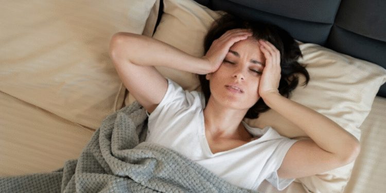 Mulher deitada na cama com expressão de incômodo e mãos na cabeça, sinalizando dificuldade para dormir