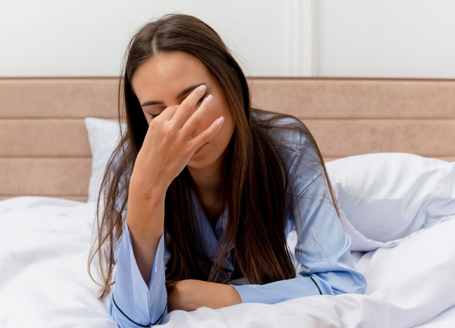 Mulher com pijama azul sentada na cama, cobrindo o rosto com a mão, demonstrando cansaço e noite mal dormida