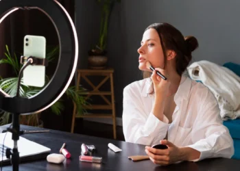Jovem aplicando maquiagem em frente a um ring light e smartphone, mostrando um tutorial de beleza, ilustrando erros de maquiagem que podem envelhecer.