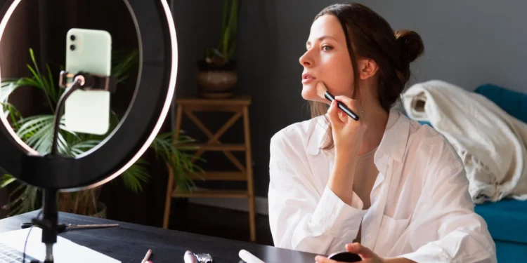 Jovem aplicando maquiagem em frente a um ring light e smartphone, mostrando um tutorial de beleza, ilustrando erros de maquiagem que podem envelhecer.