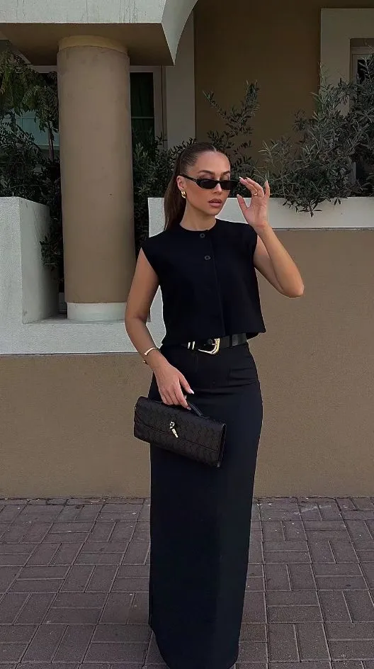 Mulher estilosa usando um look preto, com blusa preta, saia longa preta, acessórios dourados e óculos de sol, posando em ambiente externo.