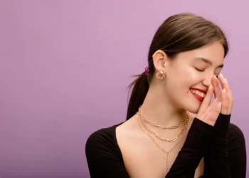 Jovem sorrindo, com acessórios dourados, incluindo brincos e colares, em fundo roxo.