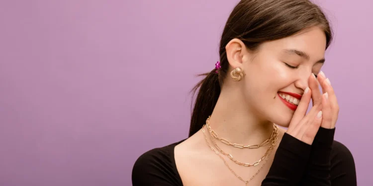 Acessórios certos mudam tudo: aprenda a dar um up no look 1 Jovem sorrindo, com acessórios dourados, incluindo brincos e colares, em fundo roxo.