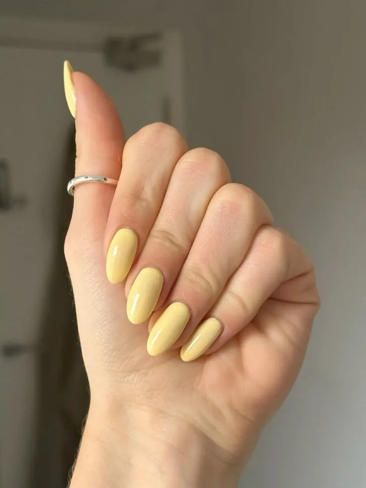 Unhas com esmalte amarelo manteiga, estilo moderno e brilhante, em formato oval.