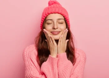 Mulher sorrindo e tocando o rosto com ambas as mãos, vestindo um suéter e um chapéu rosa, demonstrando satisfação no frio.