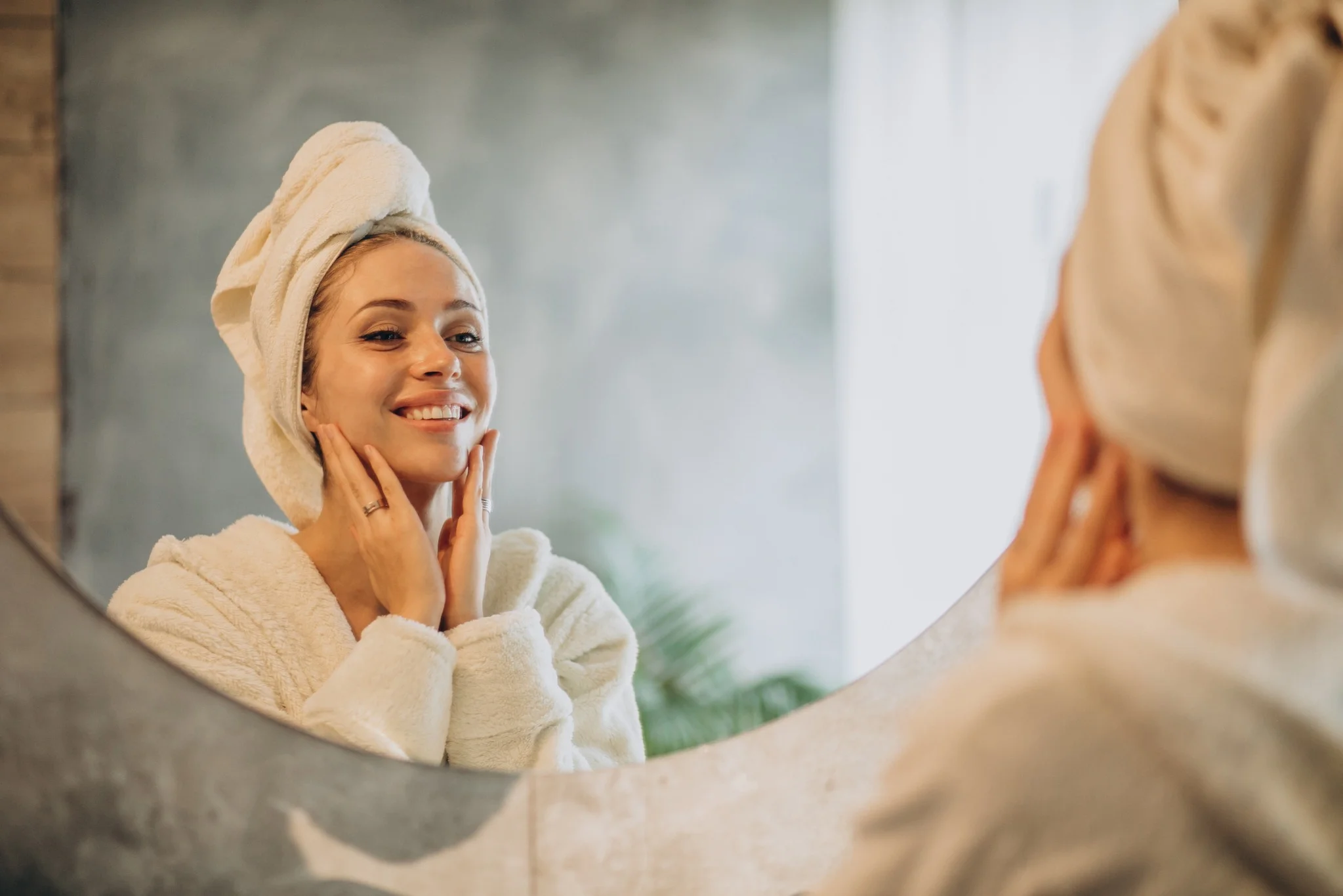 Mulher sorrindo enquanto cuida da pele, aplicando um produto no rosto após o banho, com toalha na cabeça.