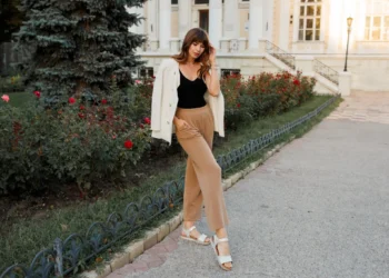 Mulher com cabelos ondulados usando uma jaqueta branca e calças largas de tom bege, posando na rua ao lado de flores, destacando o visual elegante e confortável com sandálias sem salto