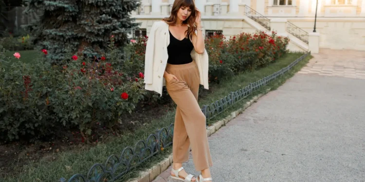 Look elegante sem salto: inspirações estilosas e confortáveis para o dia a dia 1 Mulher com cabelos ondulados usando uma jaqueta branca e calças largas de tom bege, posando na rua ao lado de flores, destacando o visual elegante e confortável com sandálias sem salto