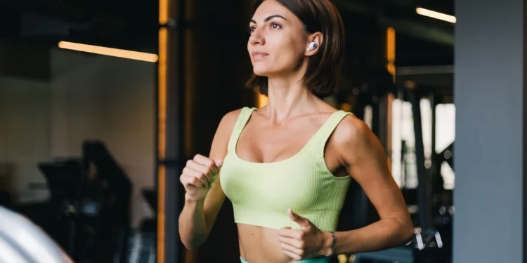 5 exercícios para aliviar os sintomas da TPM e melhorar seu bem-estar 1 Mulher correndo na esteira em academia, usando roupa esportiva verde e fones de ouvido sem fio, como parte de um exercício para aliviar os sintomas da TPM.