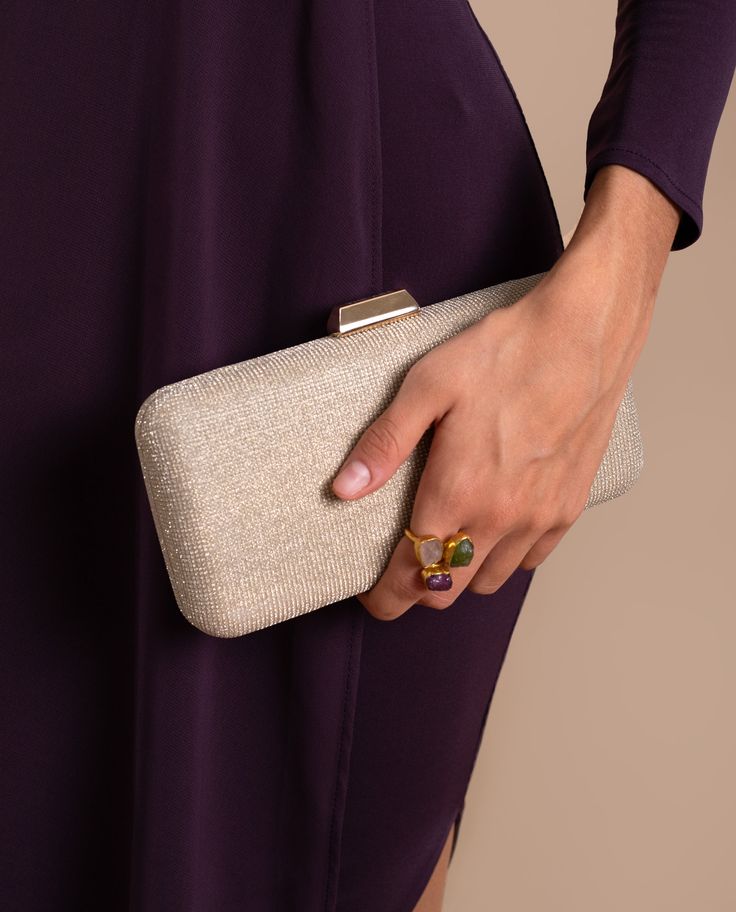Mulher segurando uma clutch dourada com vestido roxo e anéis coloridos.