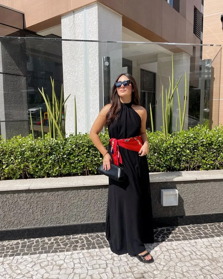 Mulher usando vestido preto e lenço vermelho na cintura, com acessórios modernos e óculos de sol, em ambiente ao ar livre.