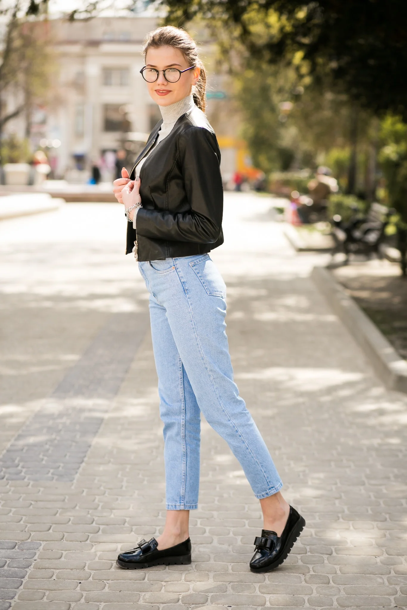 Mulher com jaqueta de couro preta, calça jeans clara e loafers pretos de salto baixo, posando ao ar livre.