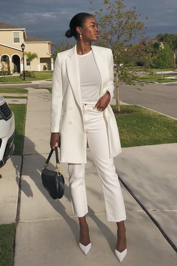 Mulher elegante vestindo um look todo branco com blazer, calça cropped e sapatos brancos, carregando uma bolsa preta.